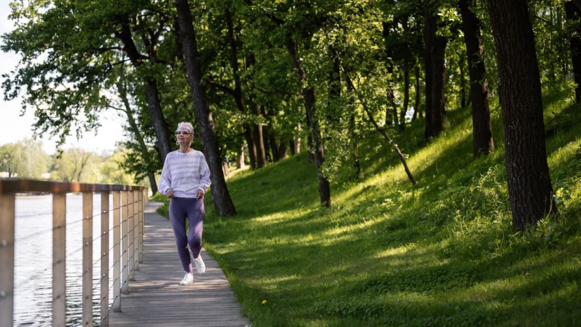Elderly woman running along a riverside path in a green park, embracing fitness and nature.